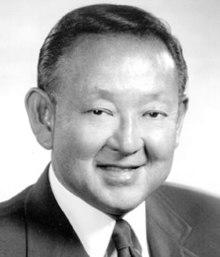 Black and white portrait of Larry Shinoda, a Japanese-American automobile designer. In the photograph, which is cropped to show his head and shoulders, Shinoda is smiling and wearing a dark suit, white shirt, and dark tie. This image was originally posted to the National Corvette Museum upon Shinoda's induction in 1998 (one year after he had died), and from appearances, may date back to the 1980s or 1990s, meaning that Shinoda appears to be in his 50s or 60s in the image. Shinoda is wearing his hair parted on the left, swept back from his forehead; the hair is almost uniformly black in the photograph.