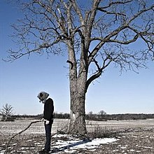 A man in an animal mask holding a stick in front of a tree