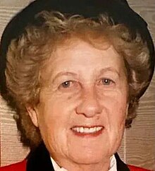 Full-face colour photograph of an elderly, smiling woman with permed blonde hair, red lipstick and a black hat on the back of her head.