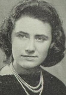 A young woman with light skin and dark curly hair, wearing a dark top and strands of pearls or beads