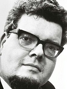 Black and white publicity shot of a bespectacled man with goatee beard, and short, curly, dark hair.