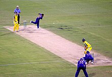 Man in a blue shirt releases a white ball with his right arm, propelling it in the air in the direction of man in yellow uniform wielding a bat at the other end of a cream piece of turf, which is surrounded by green grass. Next to the bowler is another man in yellow with a bat and a man in a light blue shirt and white hat, the umpire. Two men in blue shirts stand behind the batsman waiting for the ball to come to them.