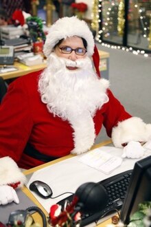 A woman in glasses wearing a red-and-white hat and fluffy coat, sitting on a desk in an office.