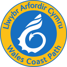 A blue circle containing a white shell stylised with a dragon tail enclosed within a yellow ring with the words Wales Coast Path in Welsh and English written on it