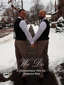 Two smiling white men wearing black vests over white shirts. They are holding hands with their bodies facing away from the viewer, though their heads are turned to face the viewer. The background is a sidewalk in winter, with snow on both sides. White text reads We Do A Documentary Film by: Rebecca Rice.