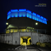 Hawley walking through a lit-up tunnel past a hooded person underneath a building with a roof illuminated in blue