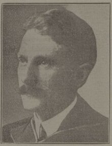 A man in early middle age, in formal academic dress (a hood just visible) and a mid-length moustache