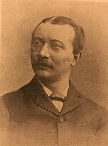 A sepia portrait photograph of a Victorian man wearing glasses, who has a moustache, and is wearing Victorian clothing, looking slightly off centre