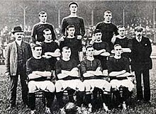 A black-and-white photo of a football team. The players are on three levels, with four on the bottom row, four on the middle row and three on the top row. The player sitting second from the left on the bottom row has a ball at his feet, and the player in the middle of the top row is wearing a flat cap.